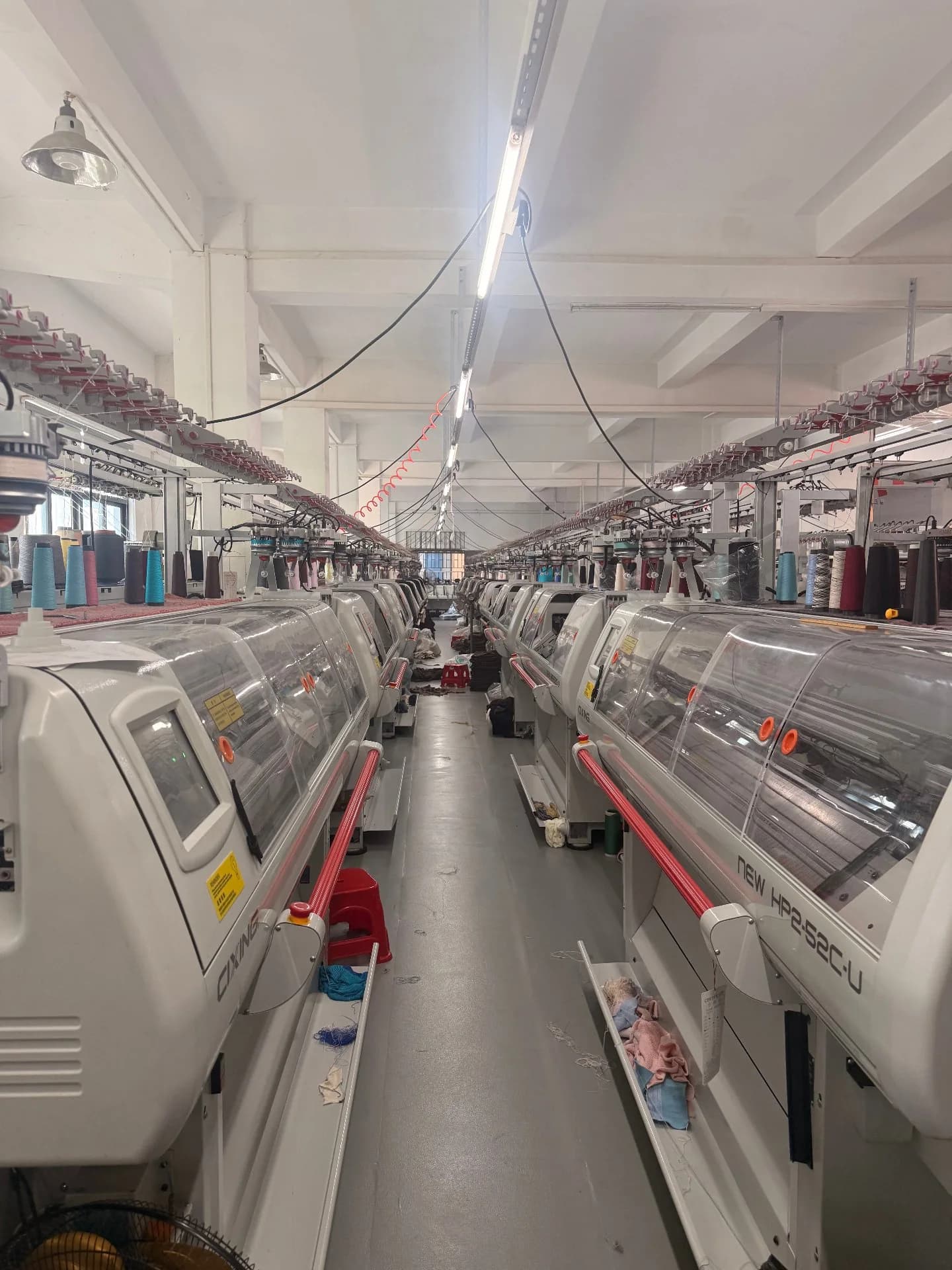 Two rows of computerised flat-bed knitting machines facing a service aisle, with a creel of yarn packages overhead and folded knitted panels in trays at the base of each machine